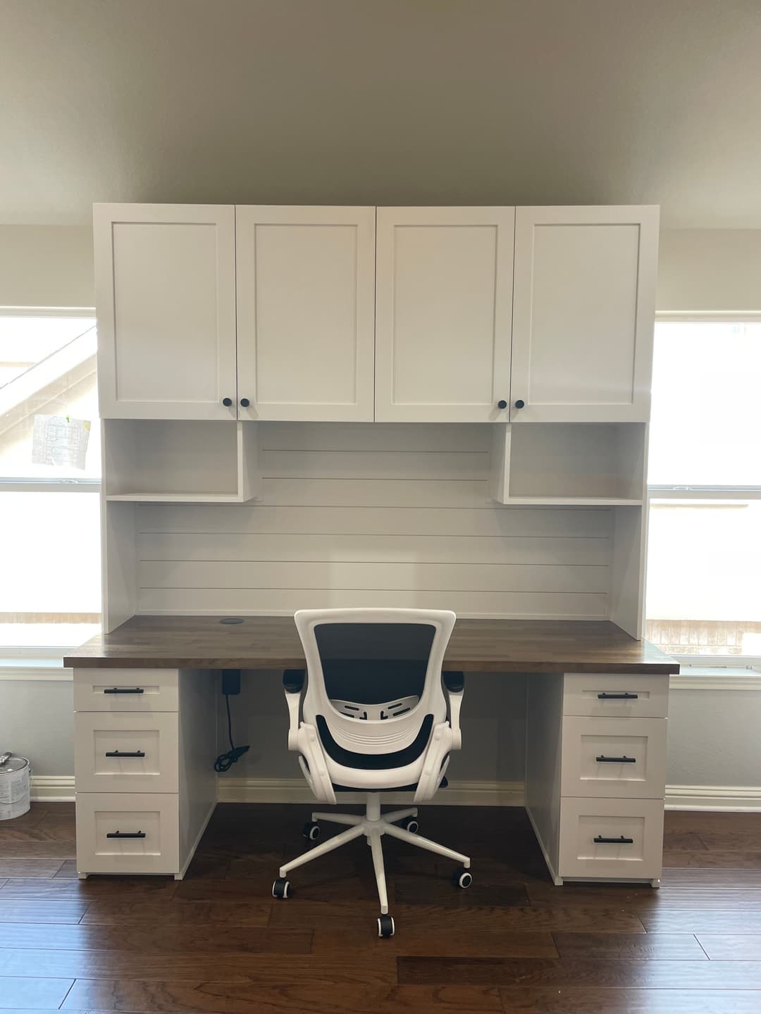 Modern white desk with upper cabinets and a rolling office chair in a bright room.